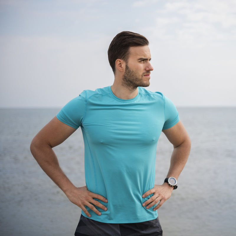 Man standing on beach in shorts and t-shirts holding water bottle.