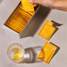 A hand is placing a yellow sachet into an open box of matching packets, with two loose packets on a light stone surface beside a glass of water. 
