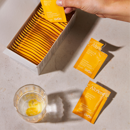 A hand is placing a yellow sachet into an open box of matching packets, with two loose packets on a light stone surface beside a glass of water. 