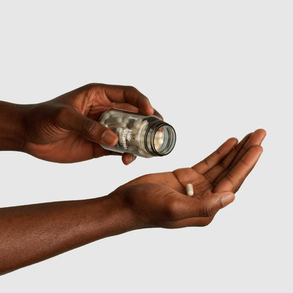 Person holding a small jar of white capsules above an open palm on a light gray background