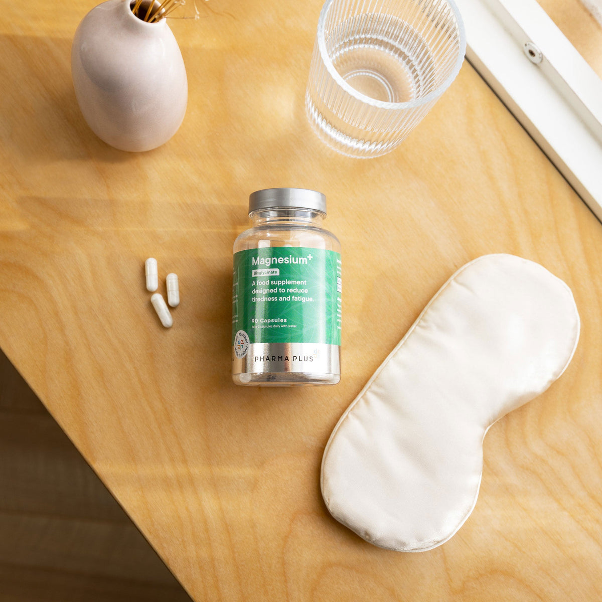 Bottle of Pharma Plus Magnesium supplement on a wooden surface with a glass of water and eye mask.