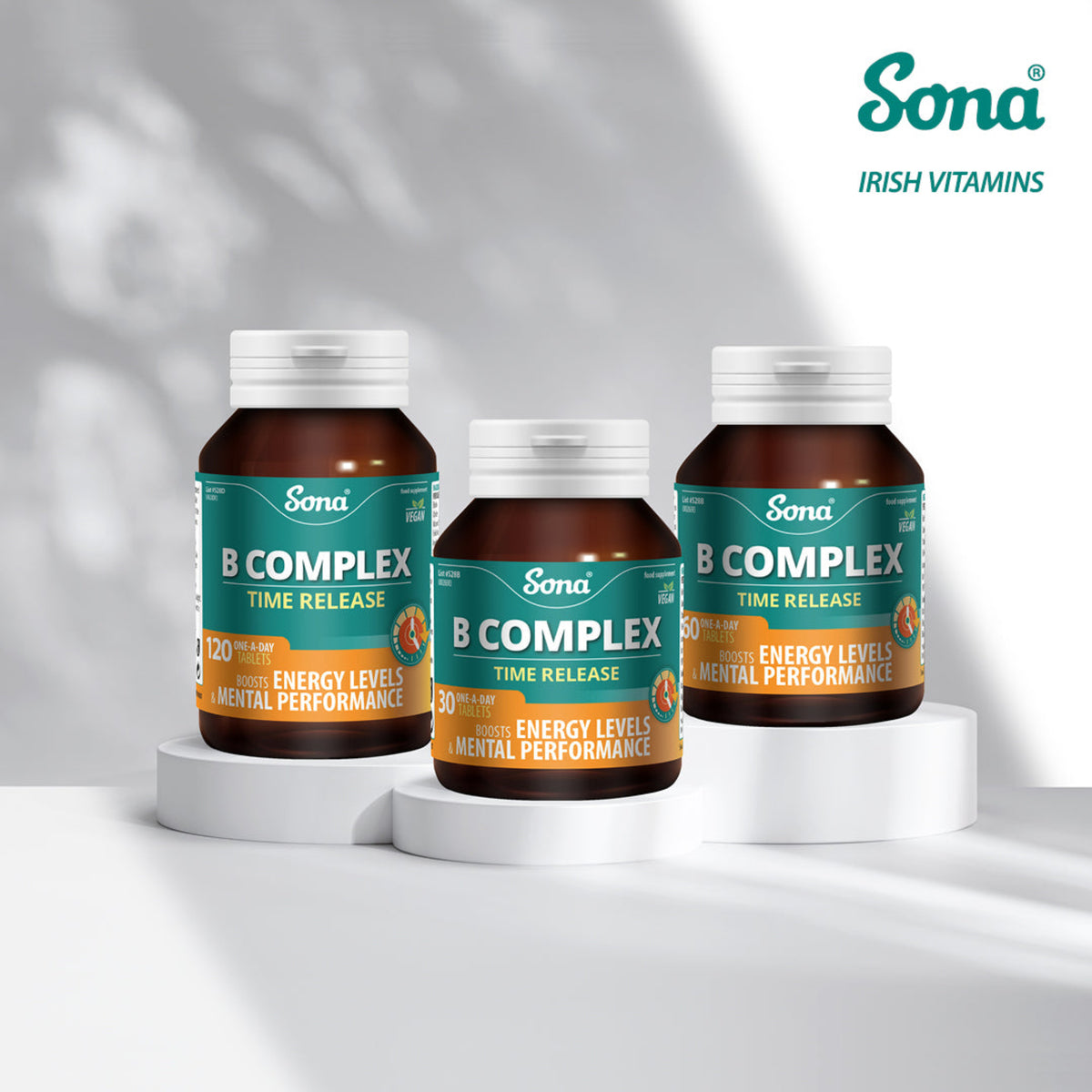 Three brown supplement bottles labeled Sona B Complex Time Release stand on white pedestals against a bright white background with soft leaf shadows. The Sona Irish Vitamins logo appears in the top right.