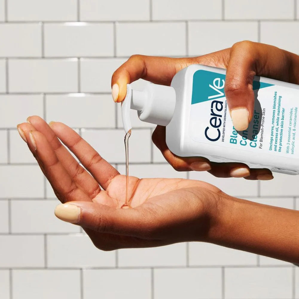 Image of transparent cleanser being dispensed from a pump bottle into the palm of a hand. Background of white tiles behind the scene.